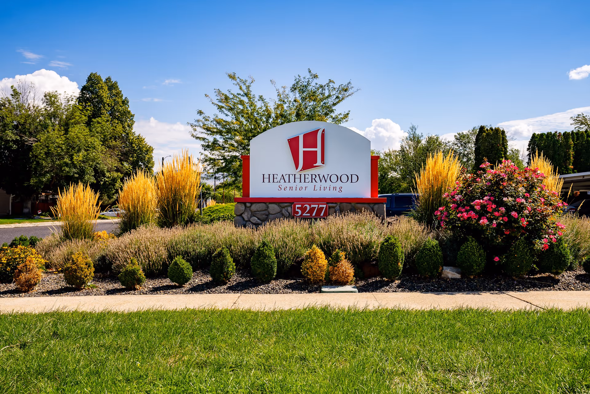 Outdoor view of a landscaped garden area with a sign for Heatherwood Senior Living, featuring the address number 5277. The sign is surrounded by various bushes, flowers, and ornamental grasses under a blue sky with some clouds.