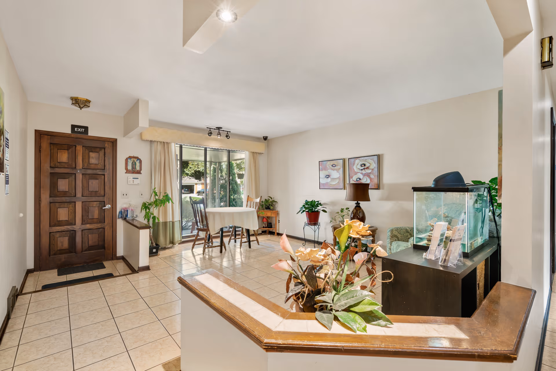 Interior view of a senior living facility common area with a wooden exit door on the left, a small table with two chairs near a large window with curtains, various plants, a fish tank on a dark wooden stand, and floral artwork on the wall.