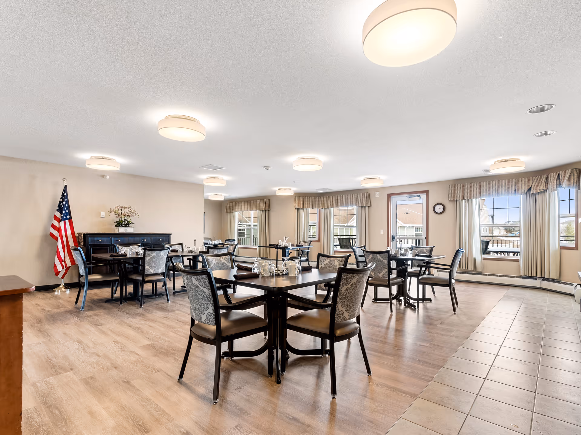 A bright and spacious dining room in a senior living facility with multiple tables and chairs arranged neatly. The room features large windows with beige curtains, allowing natural light to fill the space. An American flag stands in the corner next to a dark wooden sideboard with a floral arrangement on top. The floor is a combination of wood and tile, and round ceiling lights provide additional illumination.