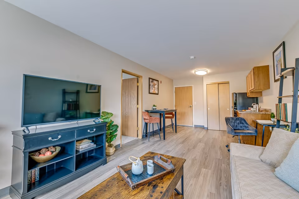 A bright and modern senior living apartment featuring a living area with a beige sofa, a wooden coffee table with a tray and cups, a black TV stand with a large flat-screen TV, and a potted plant. In the background, there is a small dining table with two chairs, a kitchenette with wooden cabinets, and a door leading to another room.