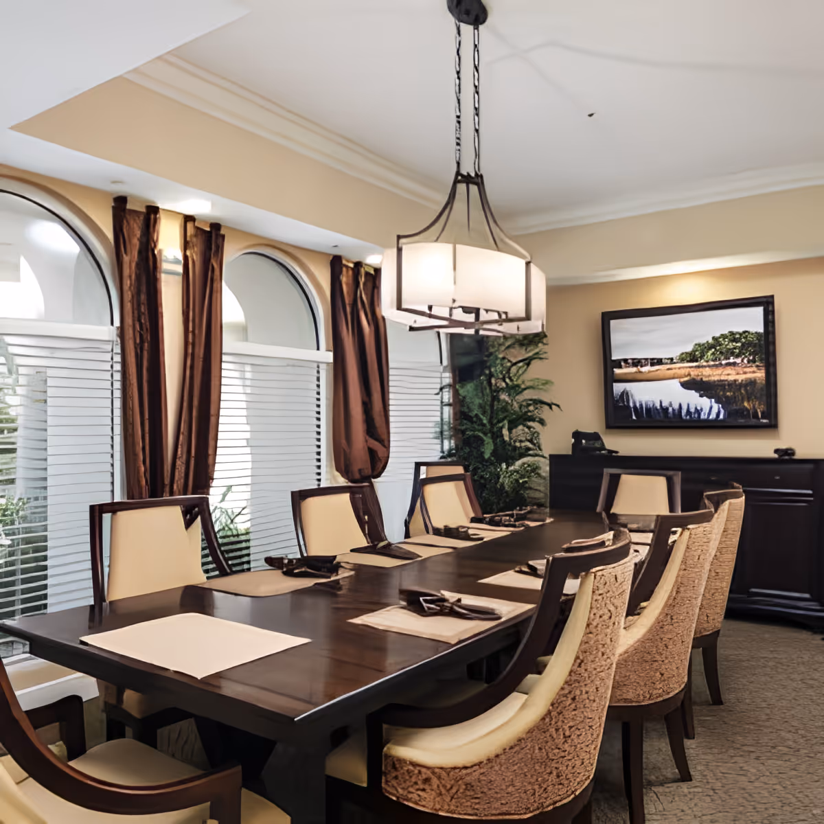 A dining room with a long dark wooden table set with placemats and napkins. The room features beige walls, large arched windows with white blinds and brown curtains, a modern chandelier hanging above the table, and a framed landscape painting on the wall. There is also a dark wooden sideboard and a green plant in the corner.