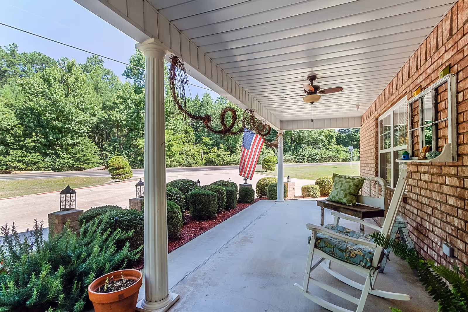 Covered front porch with rocking chairs, columns, an American flag, and landscaped shrubs.