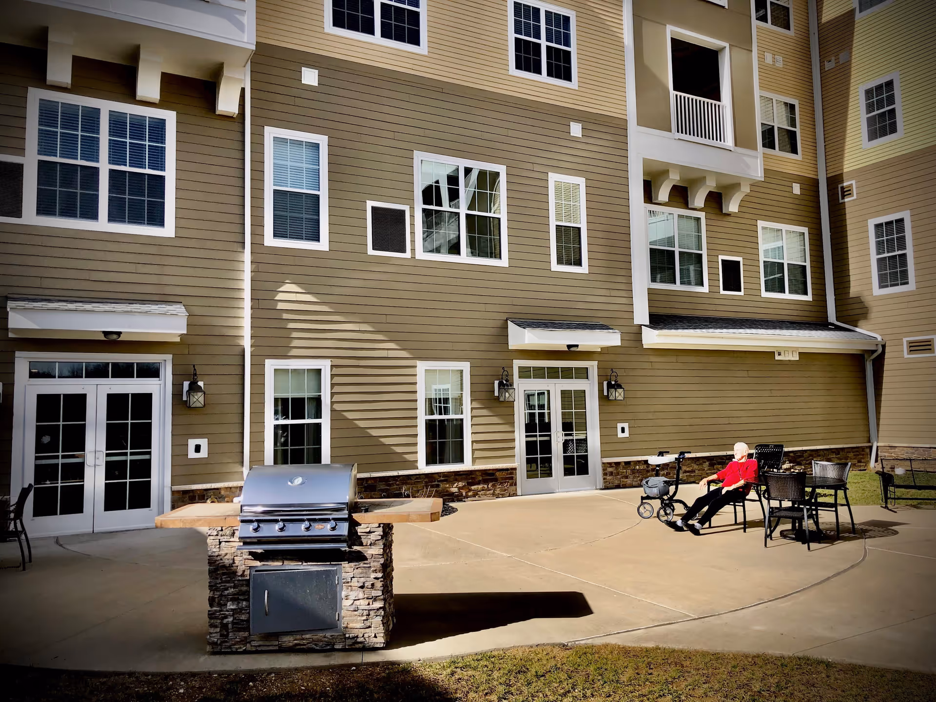 Outdoor patio beside a multi-story beige building with a stone-faced grill, patio tables and chairs, and an elderly person seated next to a walker.