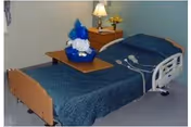 A nursing facility bedroom with a single hospital bed covered in a blue blanket, an overbed table holding a wrapped gift basket, and a bedside table with a lamp.