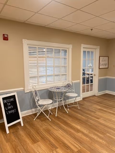 Small seating area with a round mosaic bistro table and two white metal chairs by a window and a glass door marked 'Lou's Lounge'.
