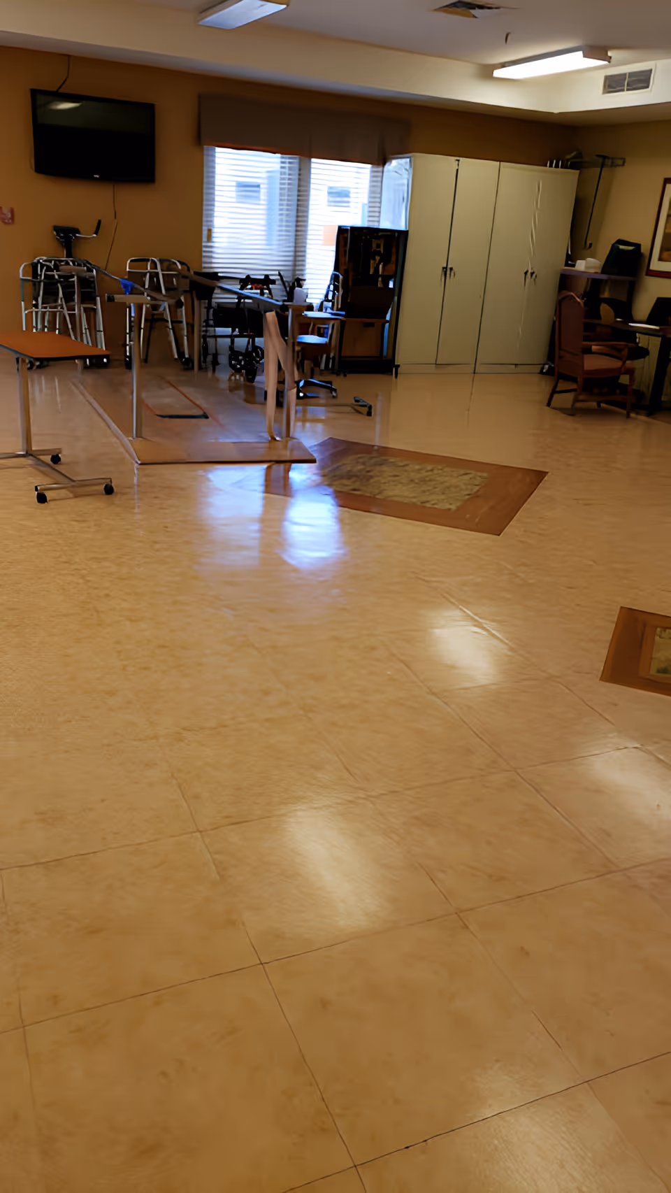 Rehabilitation/activity room with parallel bars, walkers, chairs, cabinets, and a wall-mounted TV.
