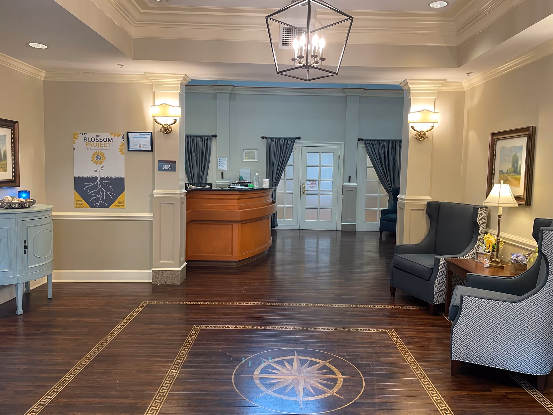 Interior view of a reception area in a senior living facility with a wooden reception desk at the center back, flanked by two columns and curtains. The floor features a decorative compass design. On the right, there are two upholstered armchairs with a small table and lamp between them. On the left, a small cabinet with decorative items and a framed poster on the wall. The lighting includes wall sconces and a modern ceiling light fixture.