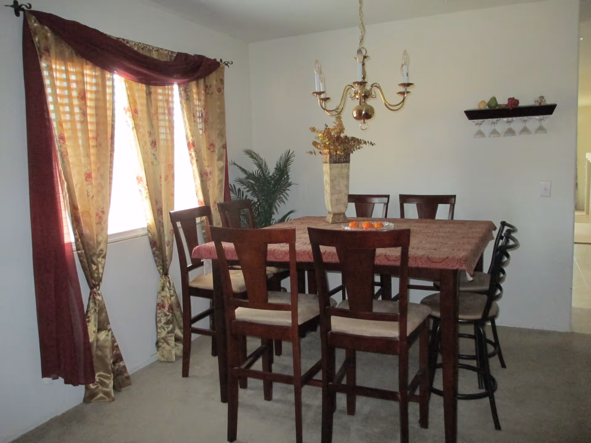 A dining room with a rectangular wooden table covered with a patterned tablecloth and surrounded by eight chairs. A decorative vase with dried flowers is on the table along with a plate of oranges. The room has beige carpet, a window with floral and burgundy curtains, a brass chandelier hanging above the table, a potted plant in the corner, and a wall-mounted shelf holding several upside-down wine glasses and decorative items.