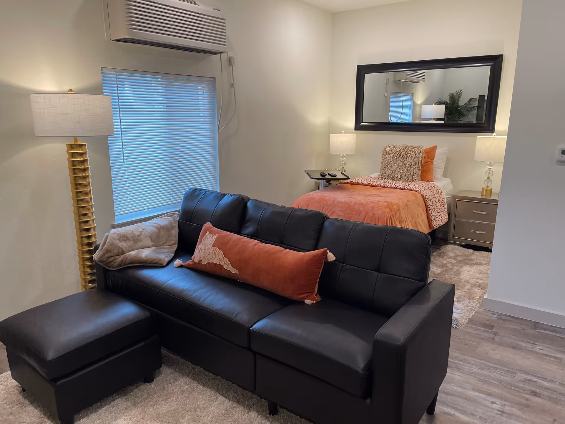 A cozy room featuring a black leather sofa with a long rust-colored pillow and a beige throw blanket. Behind the sofa is a single bed with orange bedding and decorative pillows. The room has a large mirror on the wall above the bed, two bedside tables with lamps, a window with blinds, and a tall gold floor lamp next to the sofa. The floor is covered with a light-colored rug and wood-style flooring.