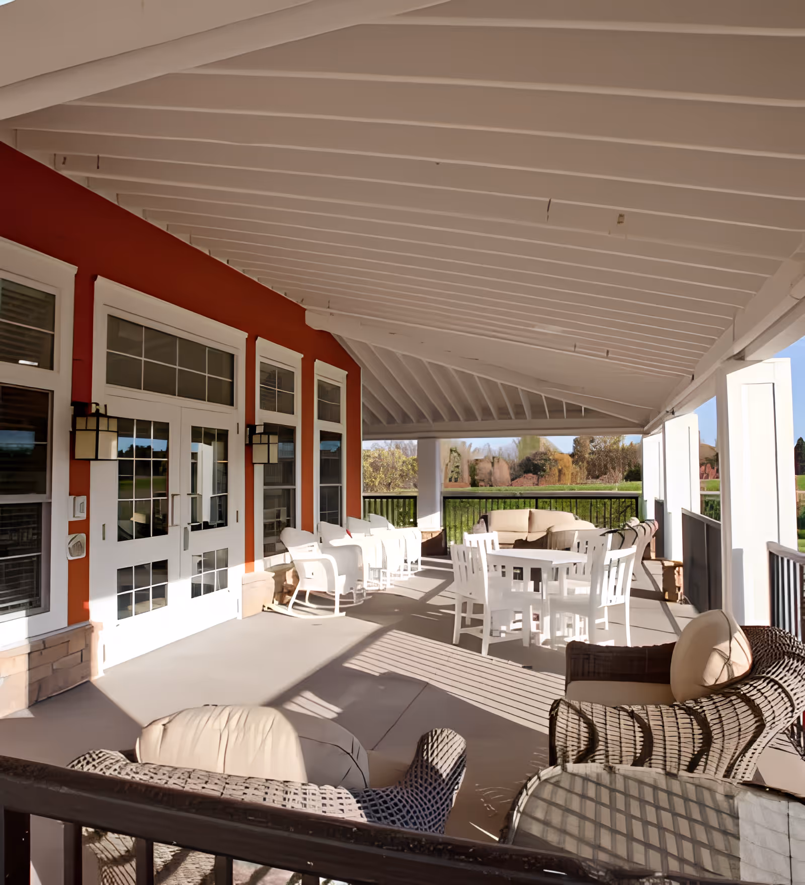 Covered outdoor patio area with white wooden ceiling and red exterior wall featuring multiple windows and doors. The patio is furnished with white chairs, a round white table, and cushioned wicker chairs. Beyond the patio railing, there is a view of green grass and trees in the background.