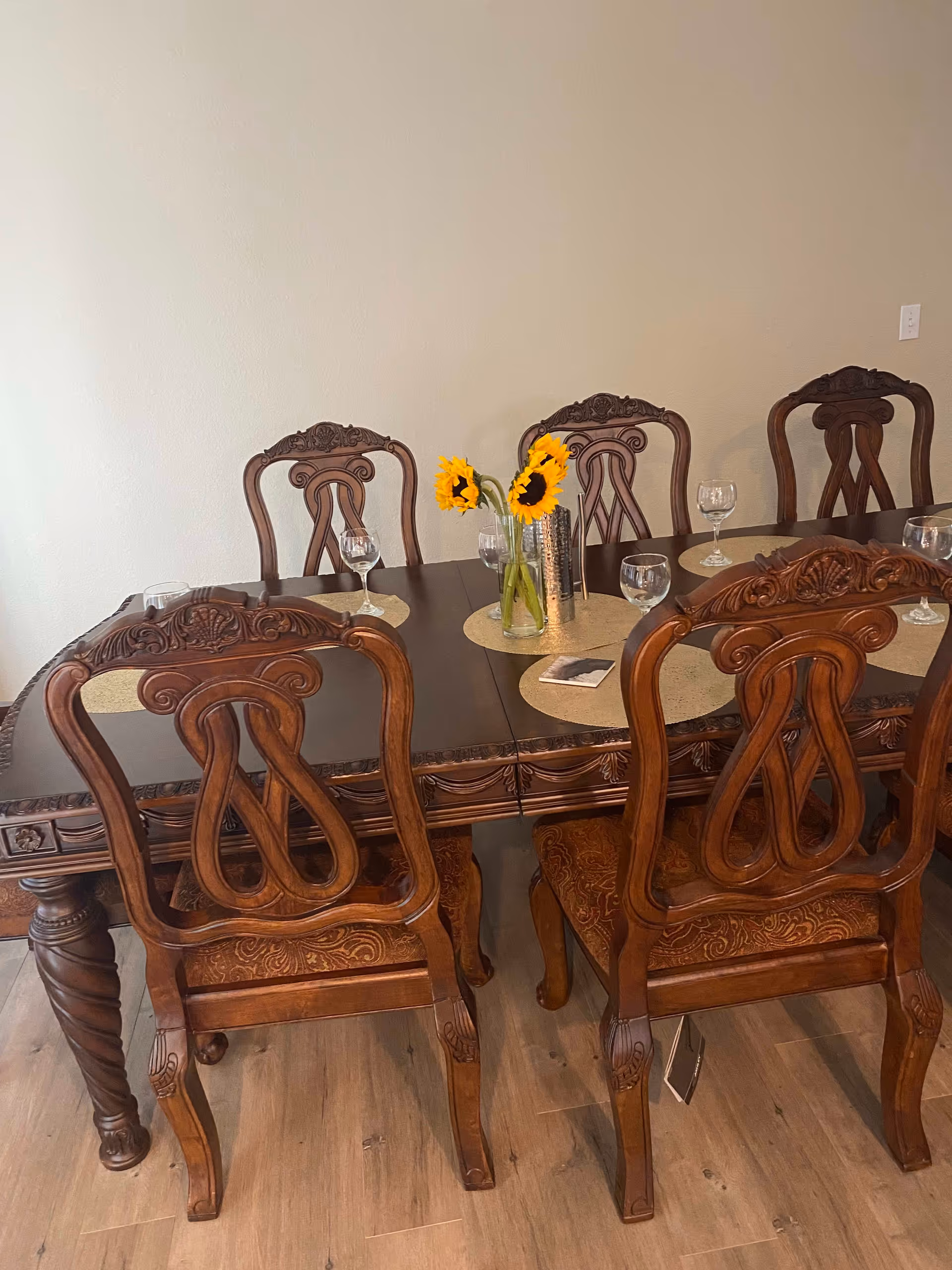 A wooden dining table with ornate carved chairs arranged around it. The table has round placemats, several empty wine glasses, and a vase with three bright yellow sunflowers. The floor is wooden, and the wall behind the table is plain and light-colored.
