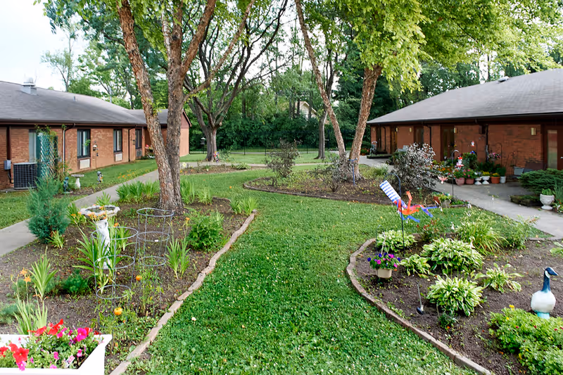 A grassy courtyard with garden beds, trees, and potted plants between two single-story brick buildings.