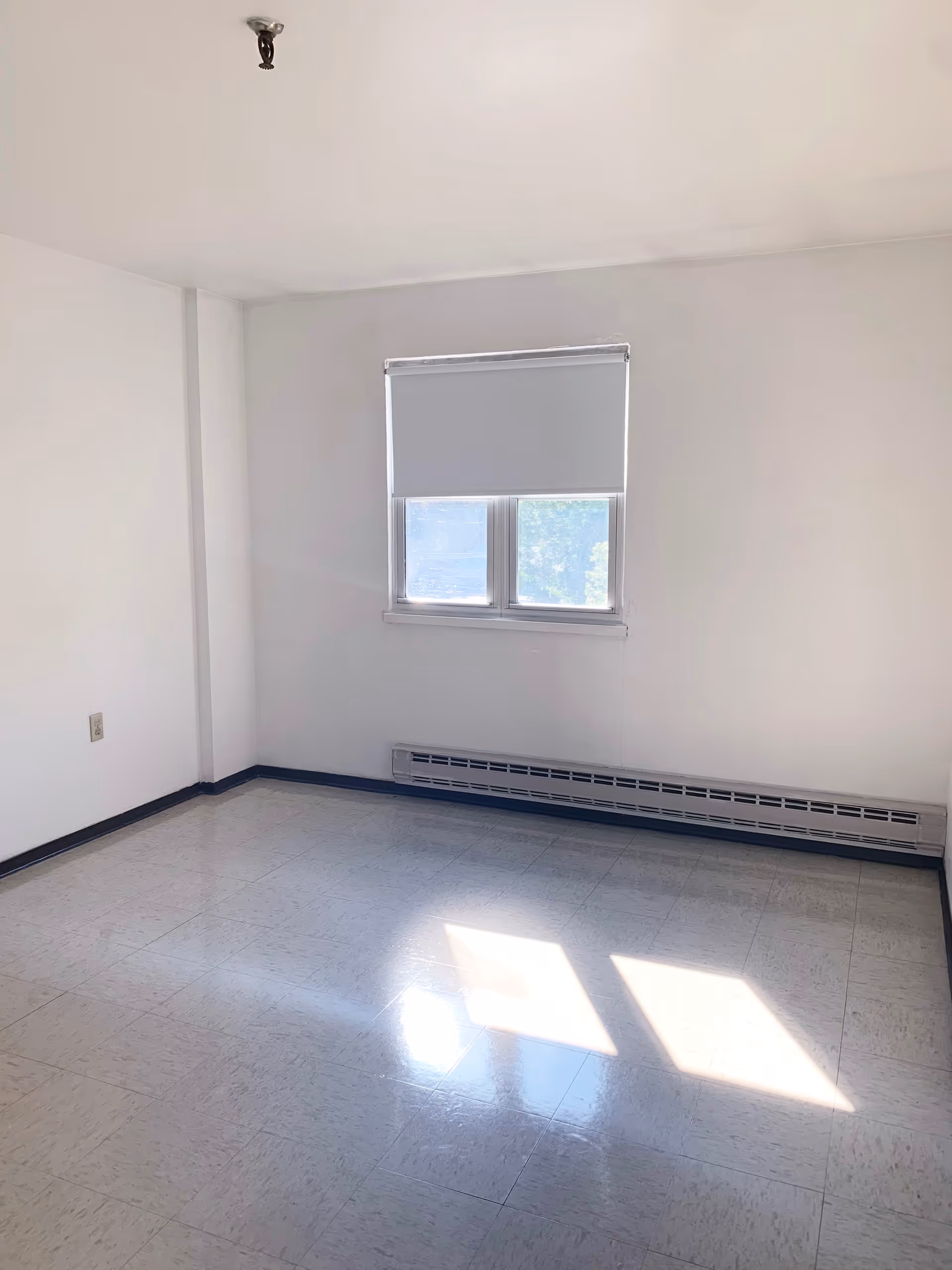 Empty room with white walls, a window with a partially closed blind, a baseboard heater below the window, and a tiled floor reflecting sunlight.