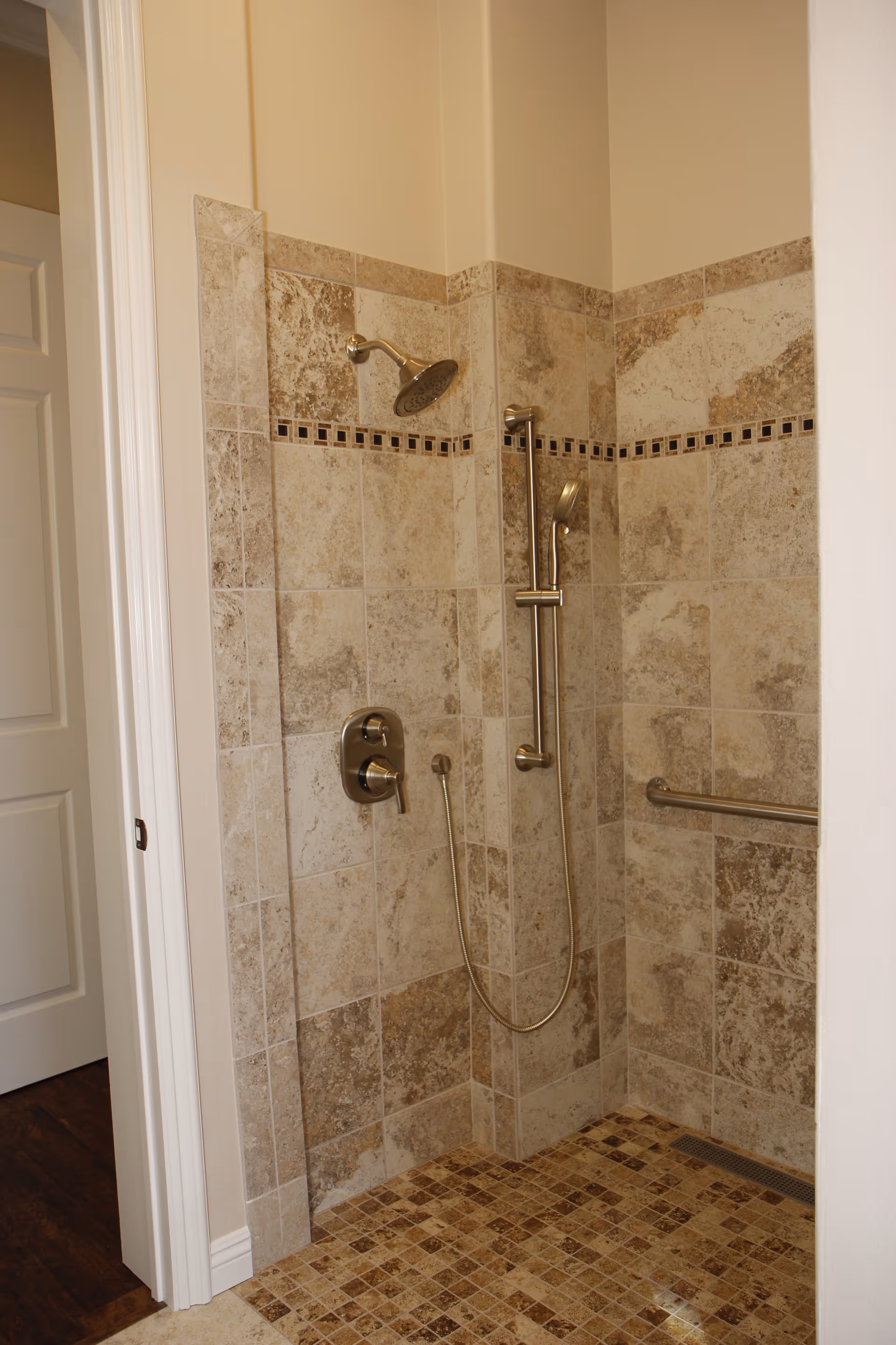 A tiled walk-in shower with beige and brown stone tiles on the walls and floor. The shower features a fixed showerhead, a handheld showerhead on a sliding bar, a control valve, and a horizontal grab bar on the wall. The shower area is open with no visible door, and a white doorframe is visible to the left.