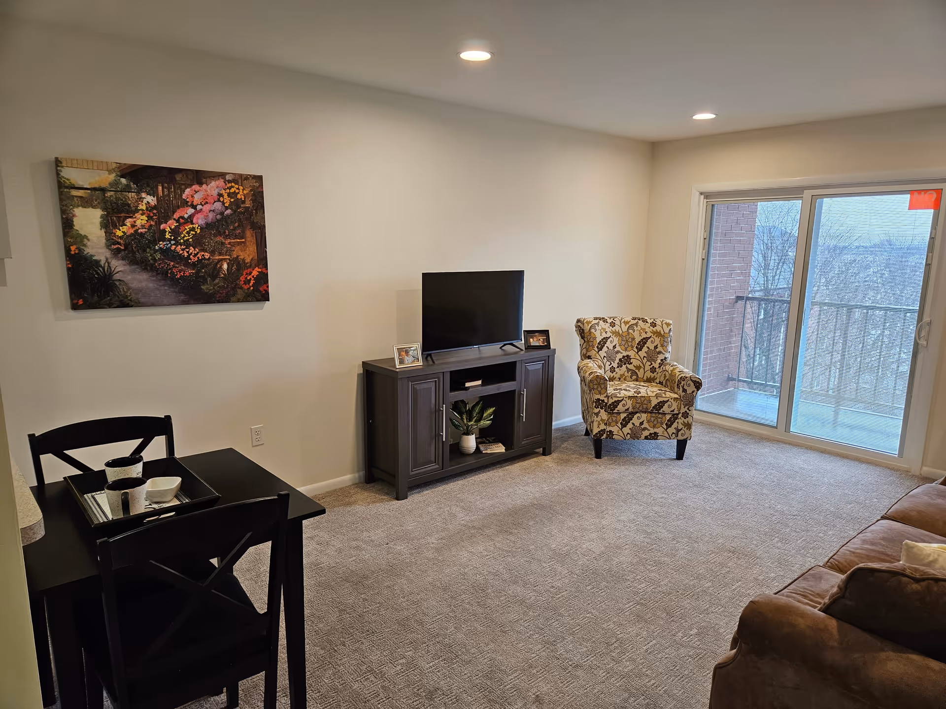 A cozy living room with beige carpet, a dark wooden TV stand holding a flat-screen TV and two small framed photos, a patterned armchair near a sliding glass door leading to a balcony, a brown sofa partially visible, a small black dining table with two chairs and a tray with cups, and a colorful floral painting on the wall.