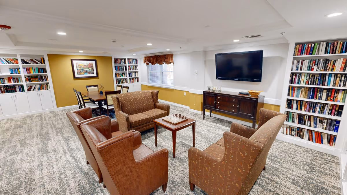 A cozy living room area in a senior living facility with two brown armchairs and two patterned sofas arranged around a small wooden table with a chessboard. The room has built-in bookshelves filled with books, a wall-mounted flat-screen TV above a wooden cabinet, a window with a brown valance, and a dining table with chairs in the background.