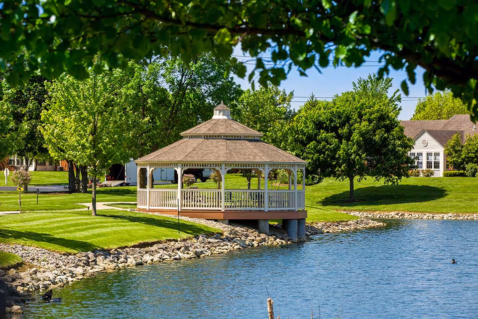 A white gazebo with a shingled roof situated on the edge of a pond, surrounded by green grass, trees, and residential buildings in the background under a clear blue sky.