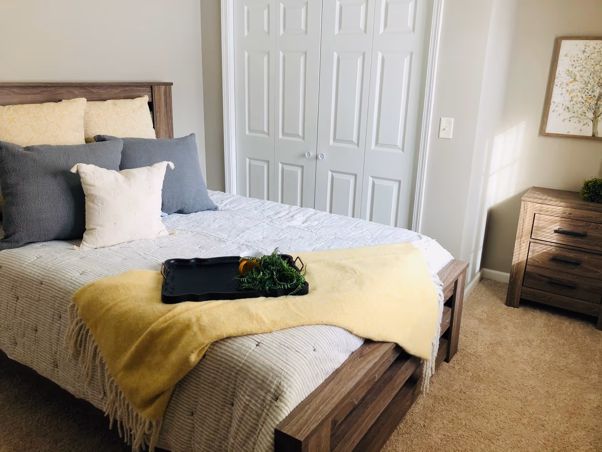 A cozy bedroom with a wooden bed frame, neatly made bed with multiple pillows in shades of yellow, gray, and white. A yellow throw blanket is draped across the foot of the bed with a black tray holding a small plant and a cup. There is a wooden nightstand with drawers on the right side, a framed picture of a tree on the wall, and white double closet doors in the background. Sunlight streams in from the right side, casting shadows on the wall.