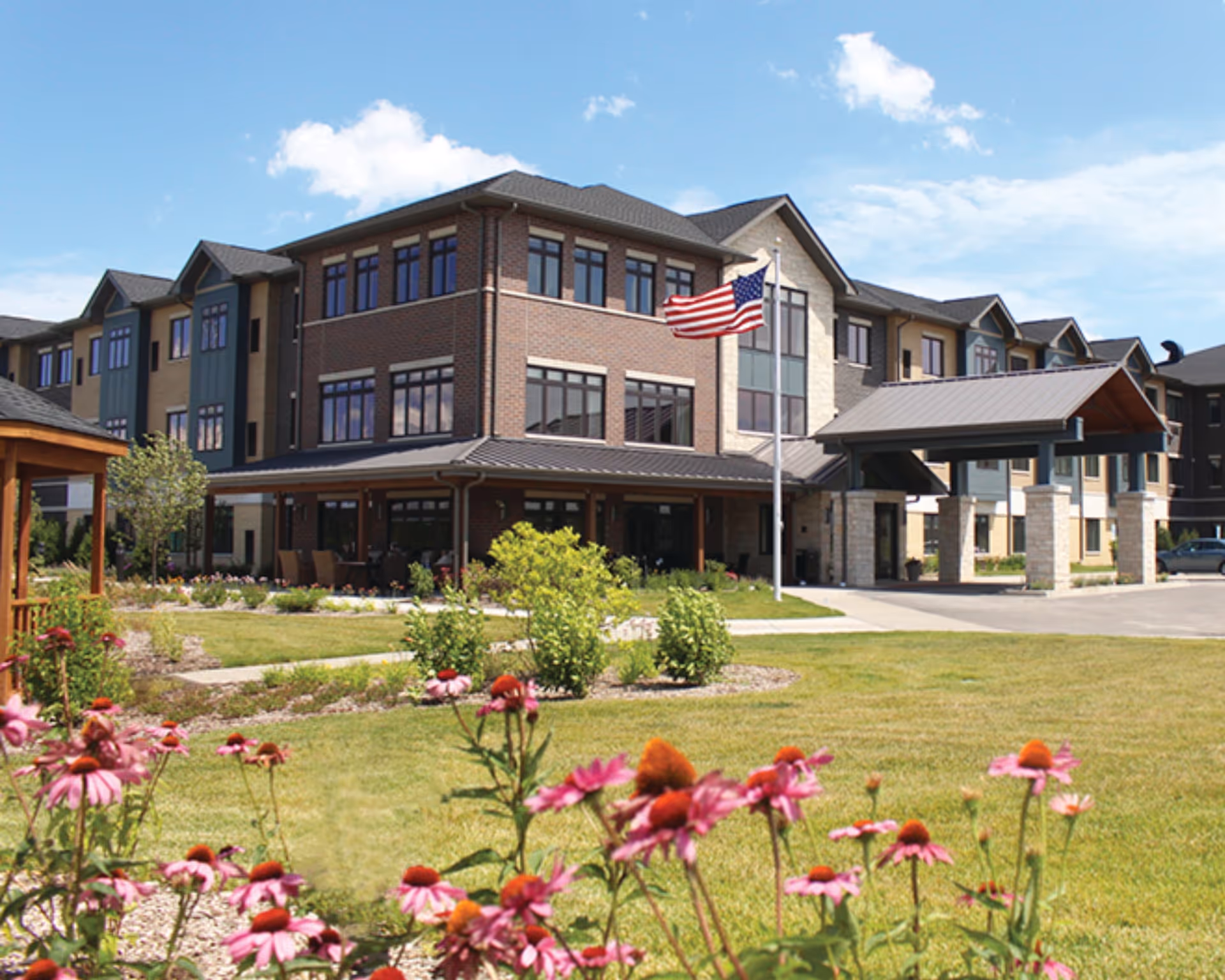 Exterior view of a three-story senior living facility building with a covered entrance, an American flag on a flagpole, landscaped gardens with green grass and pink flowers in the foreground, under a partly cloudy blue sky.