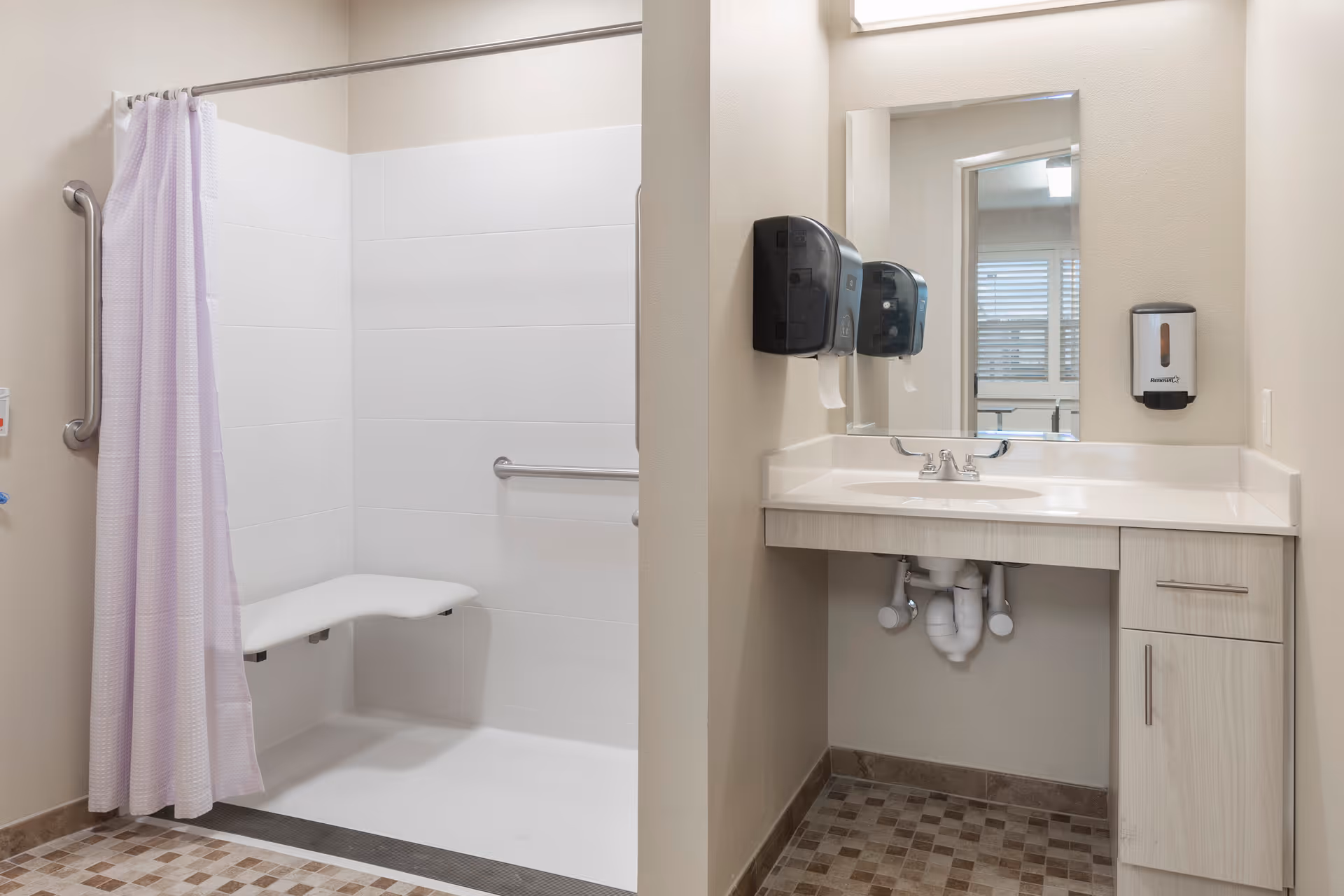 A clean and accessible bathroom featuring a walk-in shower with a fold-down seat and grab bars, a lavender shower curtain, and a vanity with a sink, mirror, and mounted soap and paper towel dispensers.