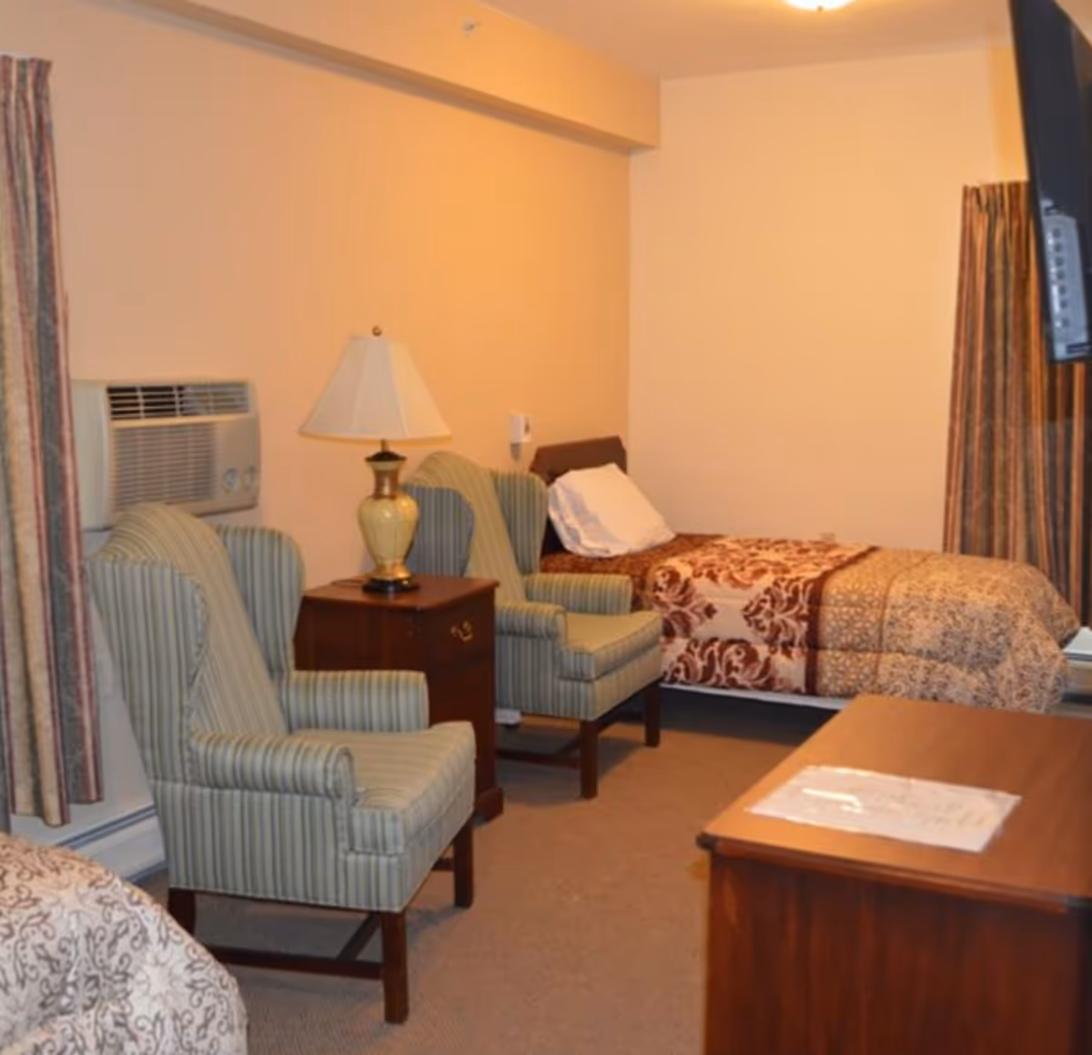 A modest bedroom with two striped armchairs, a wooden nightstand and lamp, and a single bed with patterned bedding.