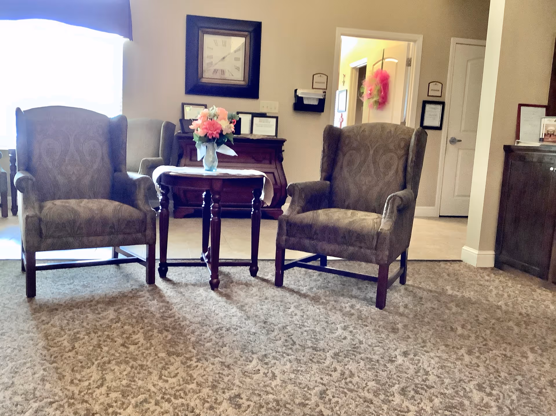 A cozy sitting area with two patterned armchairs facing a small wooden table with a vase of pink and white flowers. Behind the chairs is a wooden cabinet with framed certificates and a large wall clock above it. The room has beige walls and carpeted flooring, with a doorway leading to another room decorated with a pink wreath.