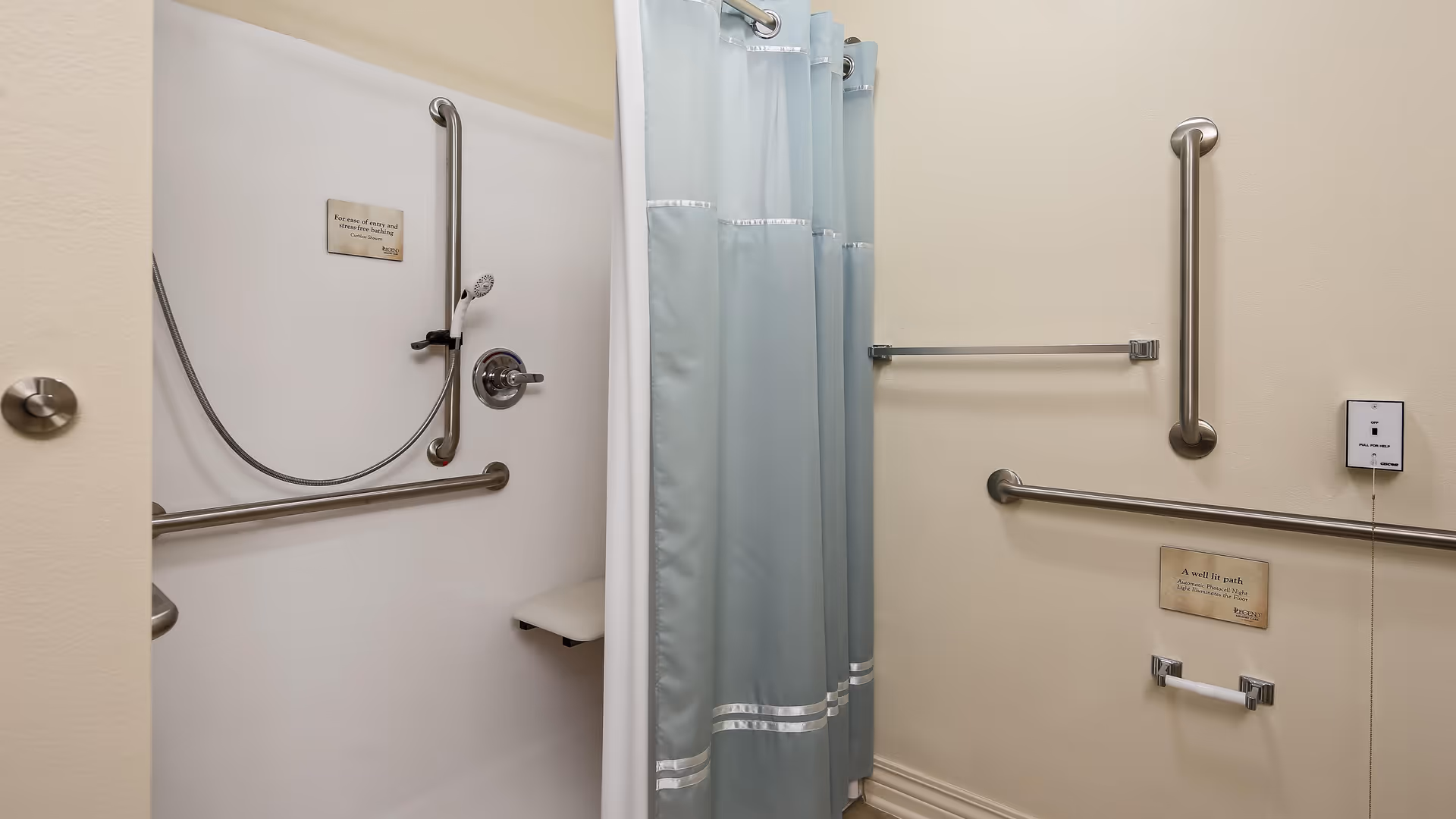 Accessible shower area with grab bars, a handheld showerhead, a fold-down seat, and a light blue shower curtain in a bathroom with beige walls.
