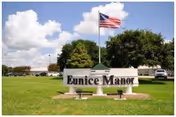 Sign reading 'Eunice Manor' on a grassy lawn with an American flag and trees behind it.