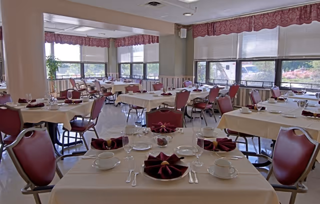 A dining room with multiple tables covered in white tablecloths, each set with plates, cups, silverware, and folded burgundy napkins. The room has large windows with blinds and burgundy valances, allowing natural light to fill the space. Red cushioned chairs surround the tables.