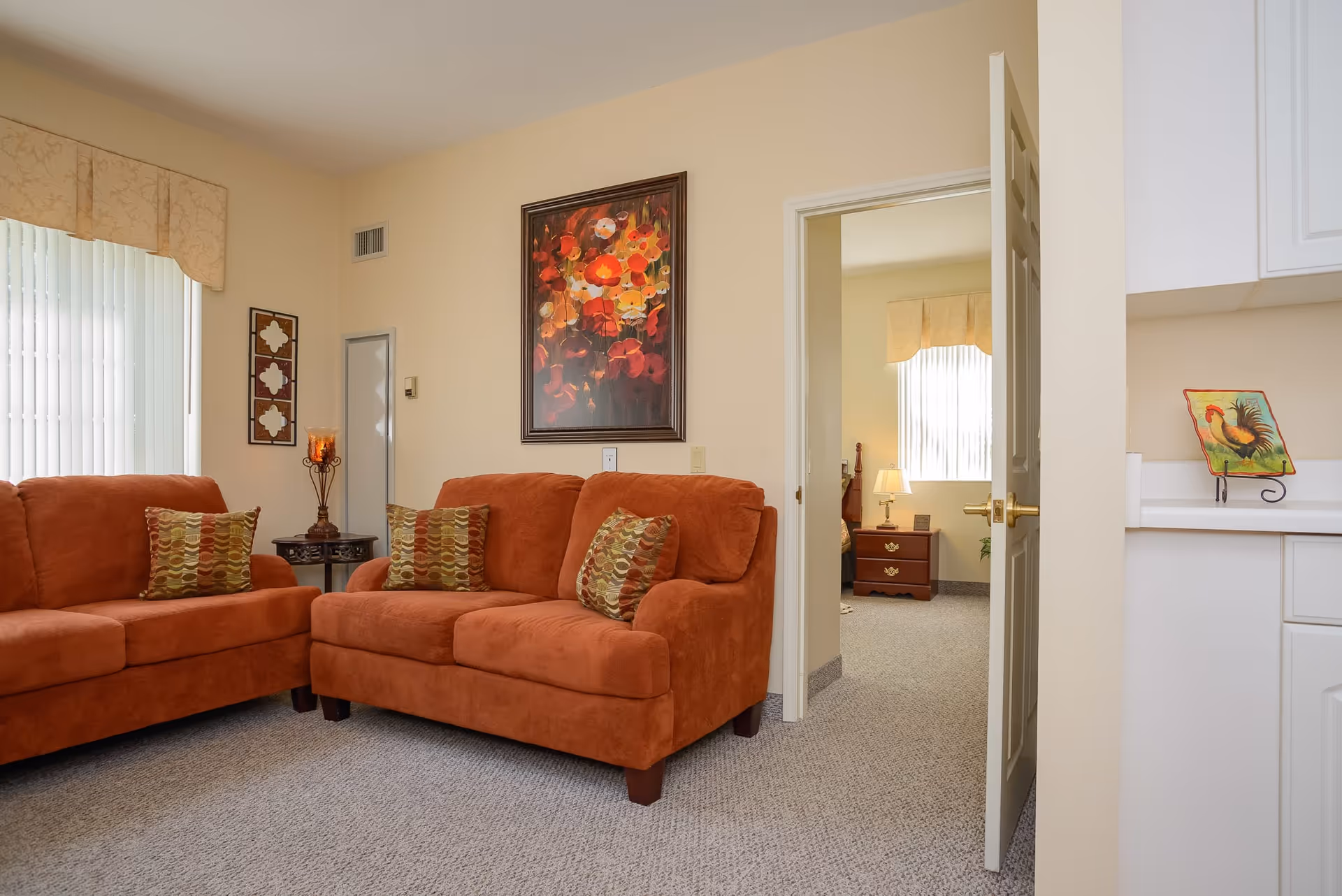 A cozy living room with two rust-colored sofas adorned with patterned cushions, a small round side table with a decorative candle holder, and a large floral painting on the wall. The room has beige walls and carpeted flooring. A doorway leads to a bedroom with a nightstand, lamp, and window with vertical blinds.