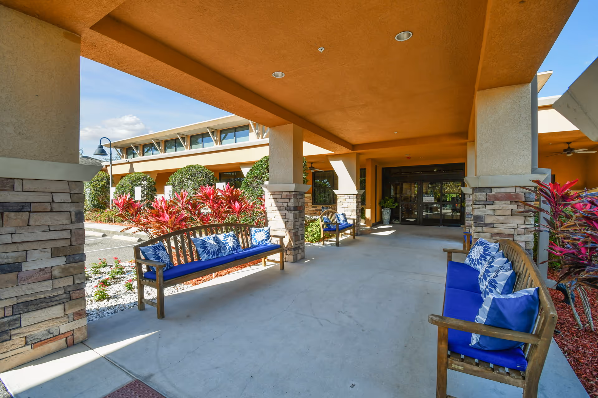 Covered outdoor seating area at the entrance of a building with wooden benches featuring blue cushions and decorative pillows. The area is supported by stone and stucco columns, with landscaping including red-leafed plants and bushes visible around the perimeter. The building has large windows and a glass entrance door under a tan-colored roof.