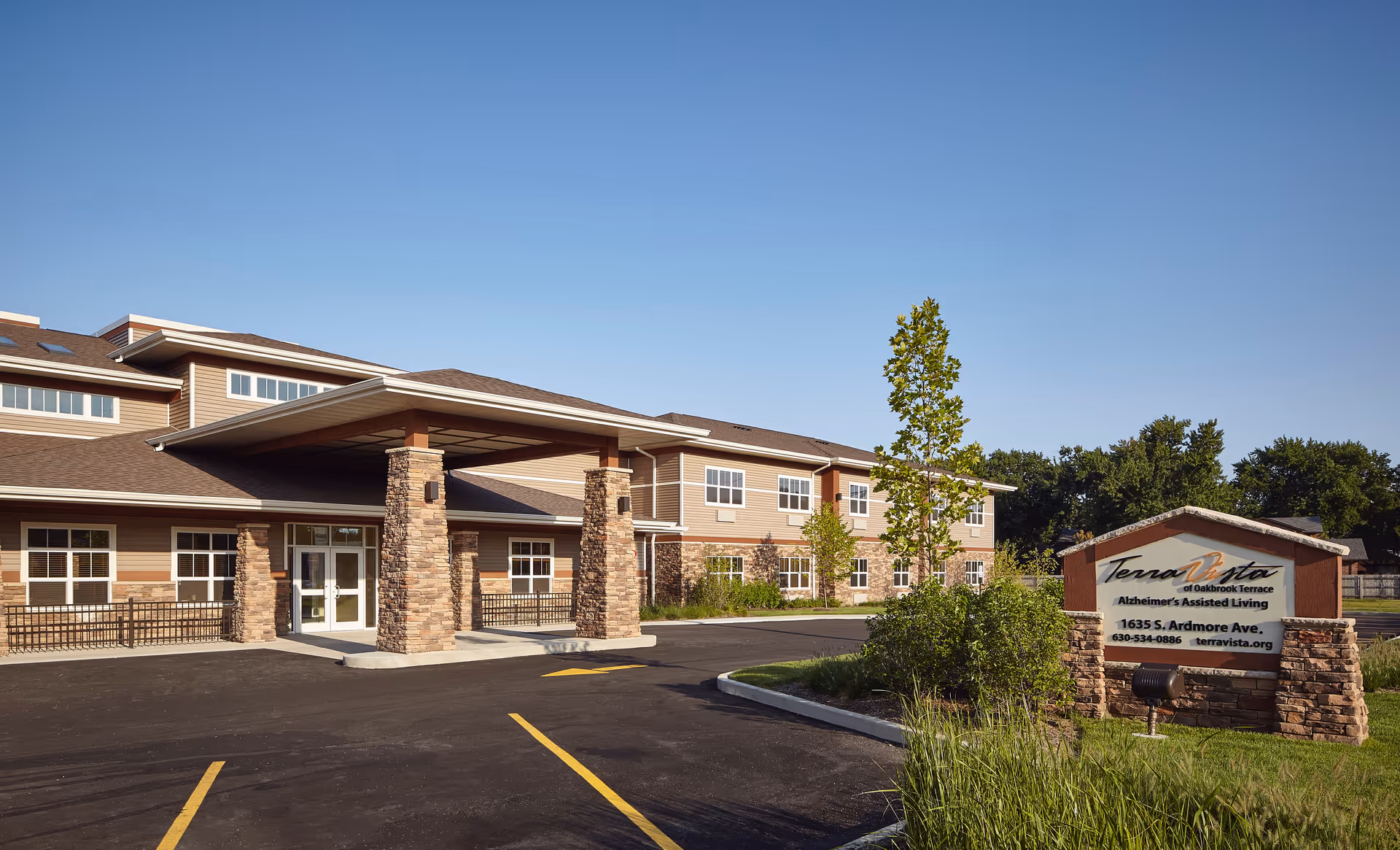 Exterior view of Terra Vista of Oakbrook Terrace, an assisted living facility. The building has a covered entrance supported by stone pillars, multiple windows, and a well-maintained parking area. There is a sign in front displaying the facility name, address, phone number, and website. The sky is clear and blue.