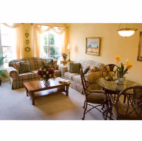 A cozy living room with two plaid sofas, a wooden coffee table with a floral arrangement, a round glass dining table with four chairs, and a window with light curtains letting in natural light. The walls are decorated with framed artwork and a wall lamp.
