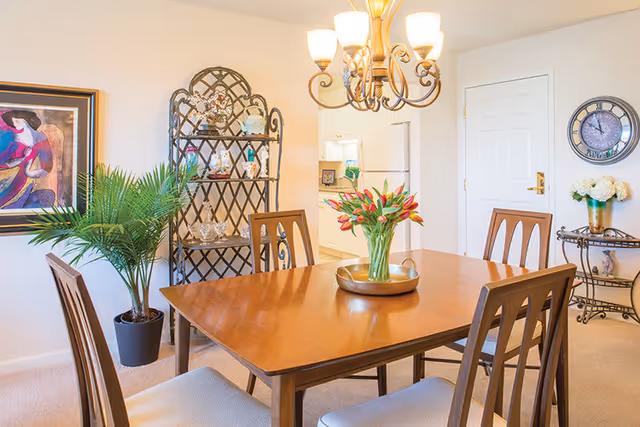 A bright dining room with a wooden table and four chairs. A vase with red tulips sits on the table. A decorative chandelier hangs above. In the background, there is a black metal shelf with decorative items, a potted plant, a framed painting on the wall, a clock, and a small table with a vase of white flowers. A white door and part of a kitchen with white cabinets and appliances are also visible.