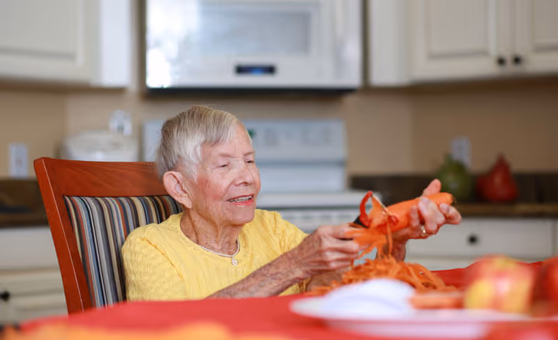 An elderly woman wearing a yellow sweater is sitting at a kitchen table peeling a carrot. The kitchen background includes a stove, microwave, and countertops with some decorative items.