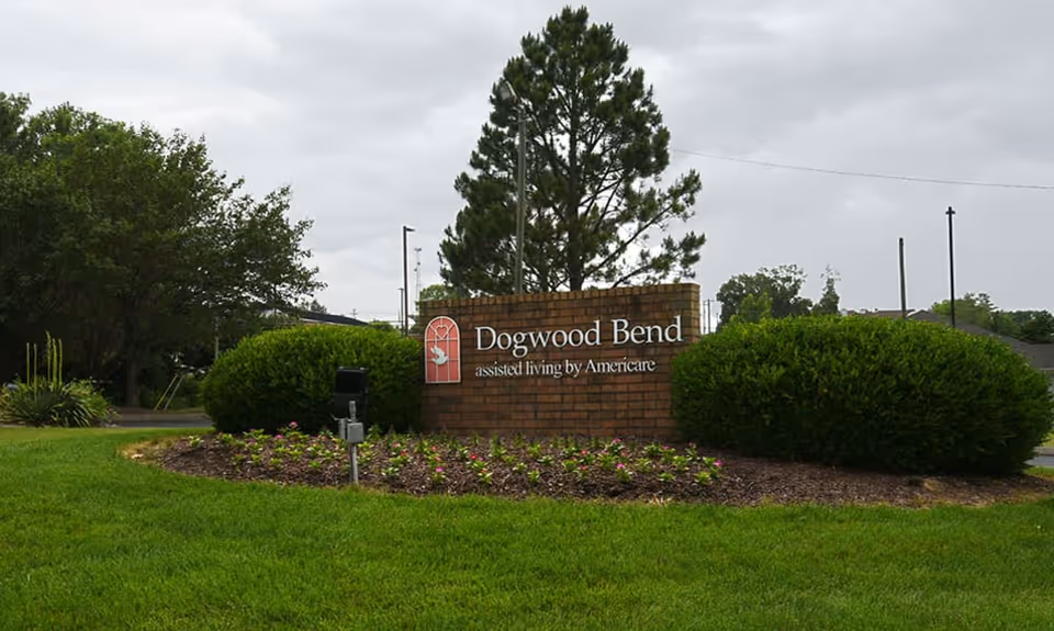 A brick sign for Dogwood Bend assisted living by Americare, surrounded by green bushes and a flower bed, with a large tree and cloudy sky in the background.