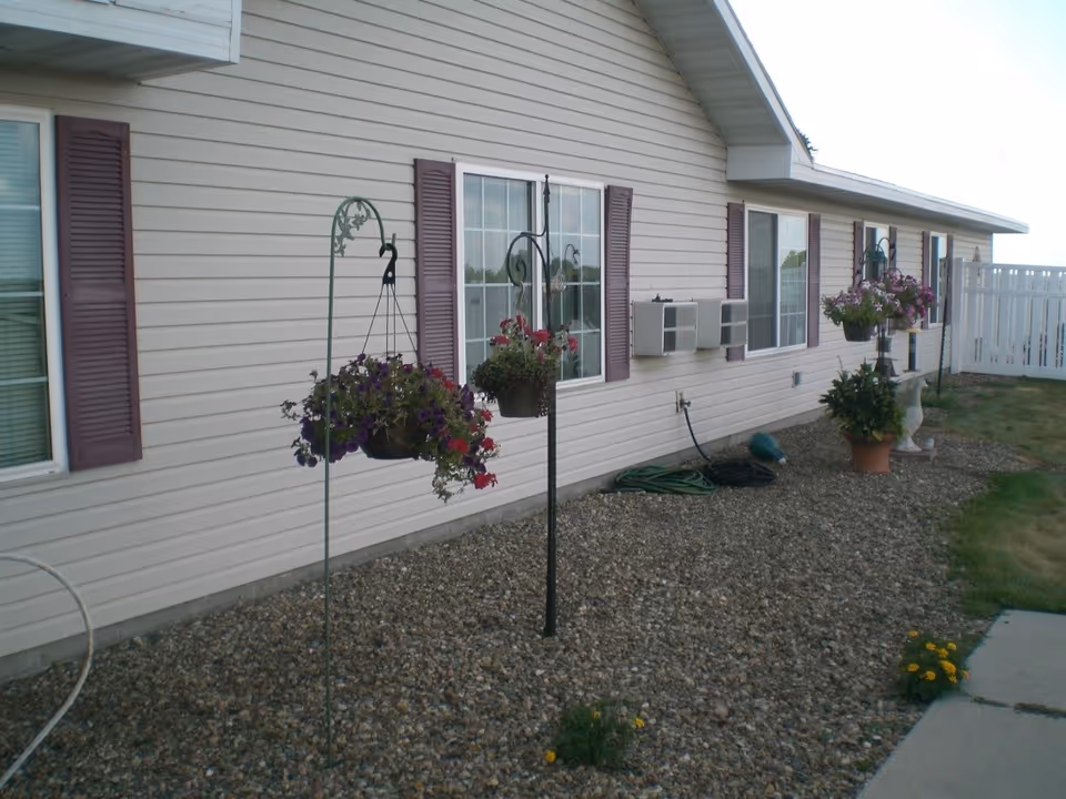 Exterior side view of a single-story building with beige siding and purple shutters. Several hanging flower baskets and potted plants are placed along a gravel bed next to the building. There are windows with air conditioning units installed, a garden hose coiled on the ground, and a white fence in the background.