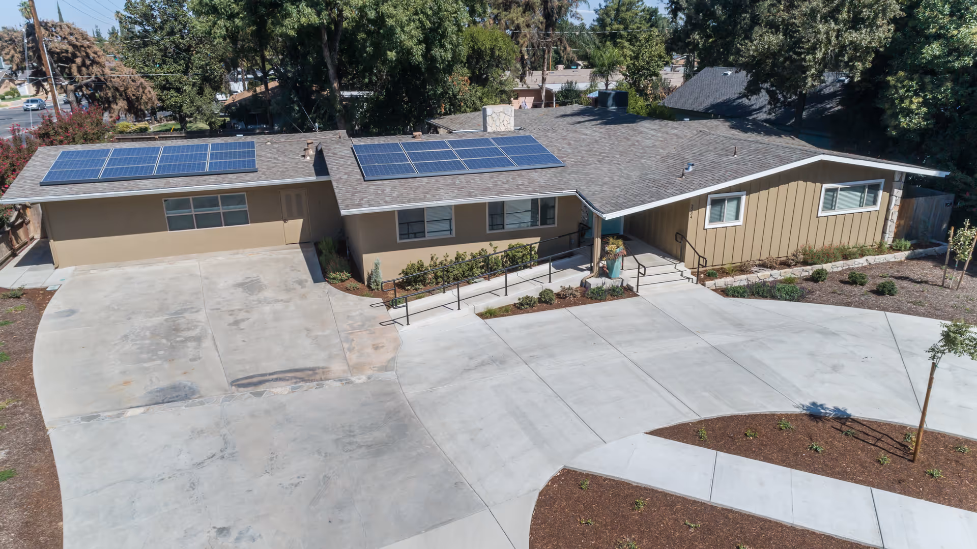A single-story building with solar panels on the roof, surrounded by trees and landscaping. The building has a beige exterior with white trim and a concrete driveway and walkway leading to the entrance, which has a covered porch and a wheelchair ramp.
