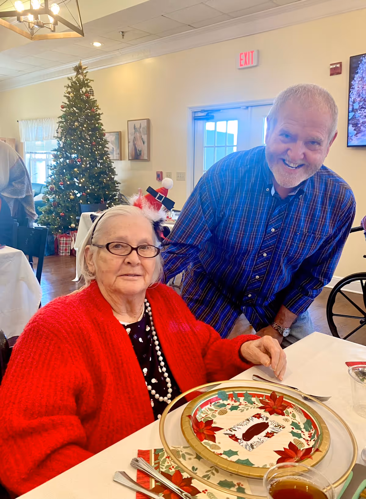 An elderly woman in a red sweater sits at a festive holiday dining table while a smiling man leans toward her and a decorated Christmas tree stands in the background.