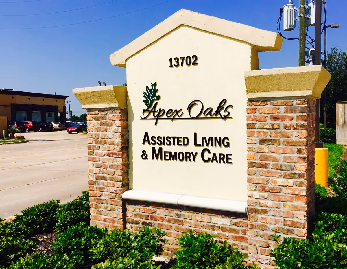 A brick and stucco monument sign at the entrance reading 'Apex Oaks Assisted Living & Memory Care' with the address 13702.