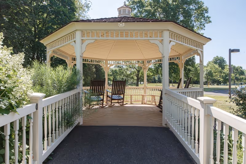 A white octagonal wooden gazebo with rocking chairs viewed from a ramp amid trees and landscaping.