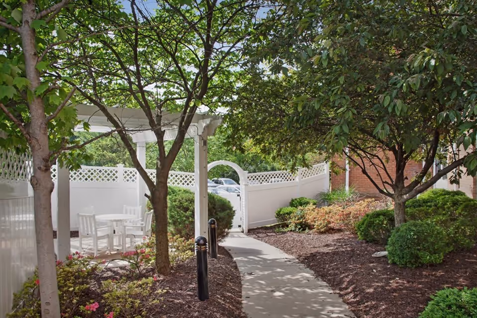 A paved walkway surrounded by landscaped bushes and trees leading to a white gate with a lattice fence. To the left, there is a white pergola with a round table and four white chairs underneath it.