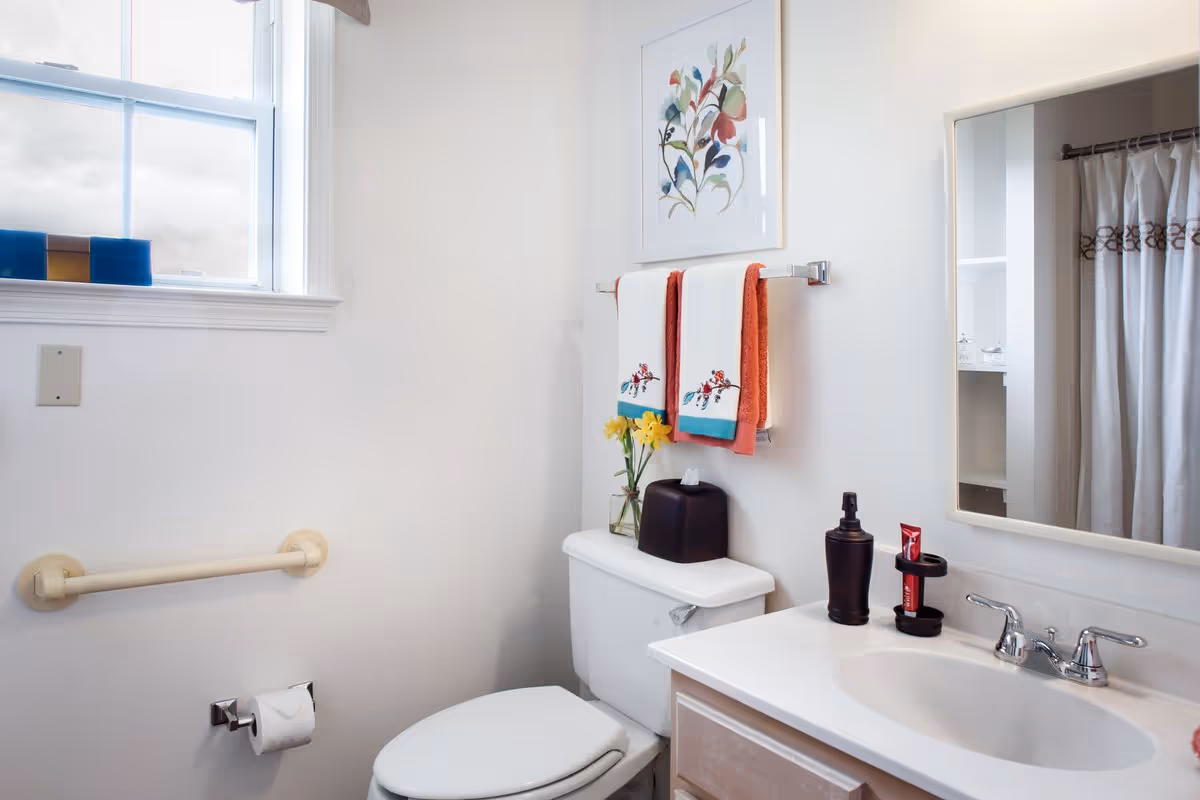 A clean bathroom with a white toilet, a white sink with silver faucet, a mirror above the sink, and a window letting in natural light. There are two decorative towels hanging on a towel rack above the toilet, a small vase with yellow flowers, a black tissue box, and soap dispenser on the sink counter. A shower curtain is partially visible in the mirror reflection.