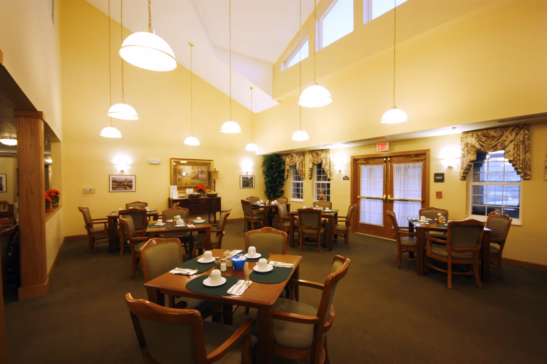 A dining room with multiple wooden tables and chairs arranged neatly. Each table is set with white cups, saucers, and silverware on green placemats. The room has high vaulted ceilings with several hanging pendant lights, yellow walls, framed artwork, and windows with patterned curtains. Double doors with exit signs are visible at the back.