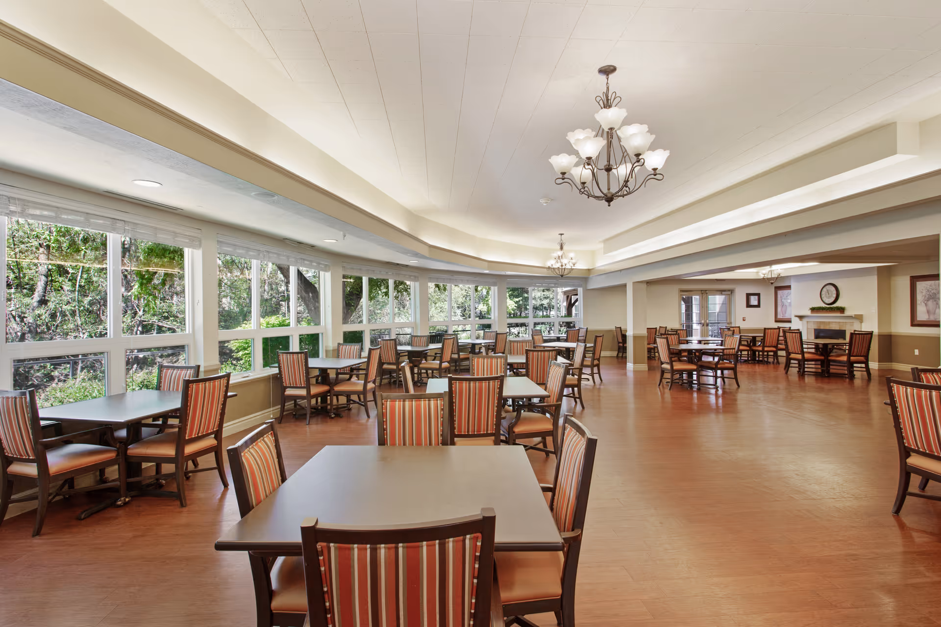 A spacious dining room with multiple tables and chairs arranged neatly. The chairs have striped upholstery and the room features large windows along one wall, allowing natural light to brighten the space. There are chandeliers hanging from the ceiling and a fireplace with a clock above it in the background.