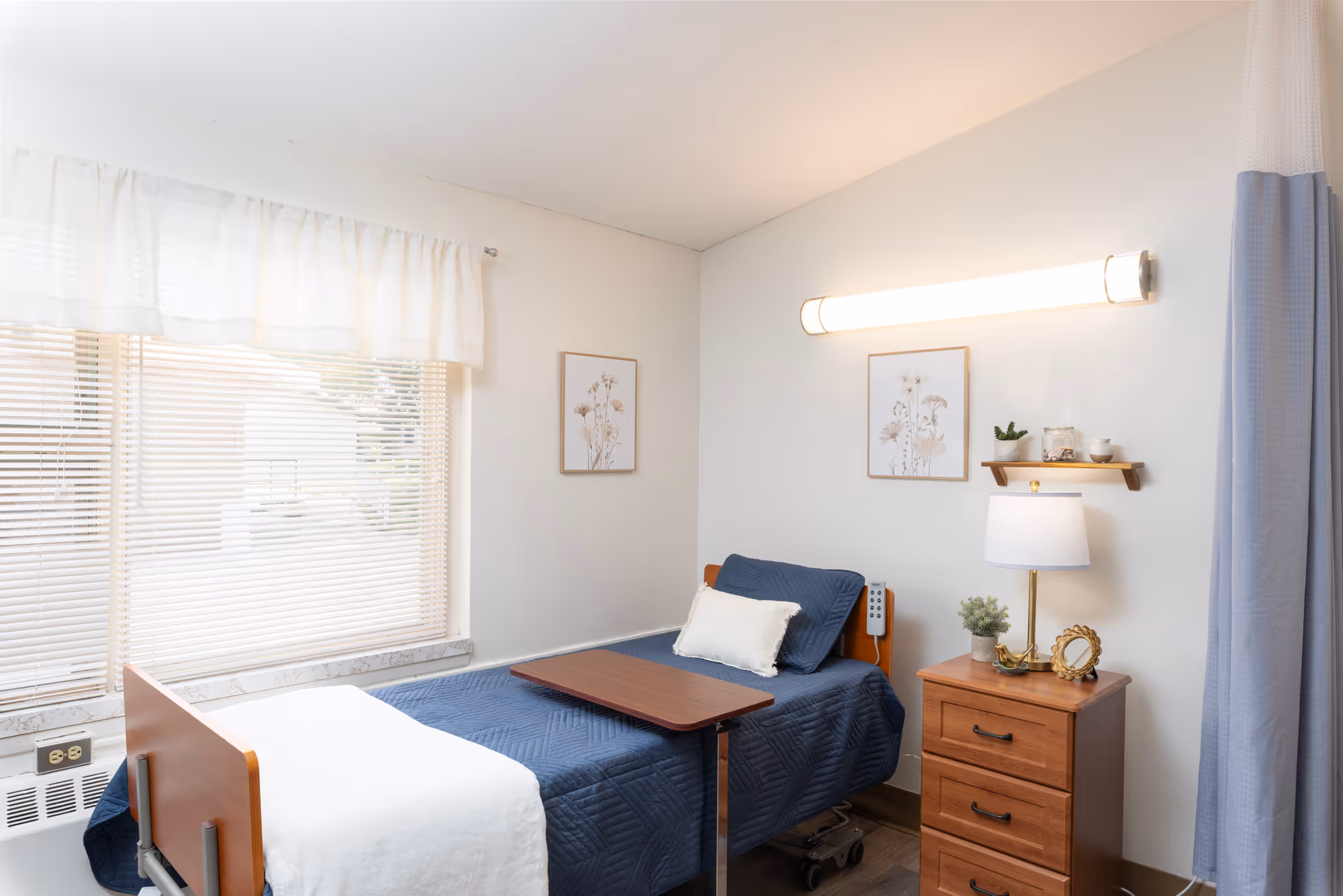 A tidy resident bedroom with a single bed, over-bed table, wooden nightstand and lamp beside a large window with blinds.