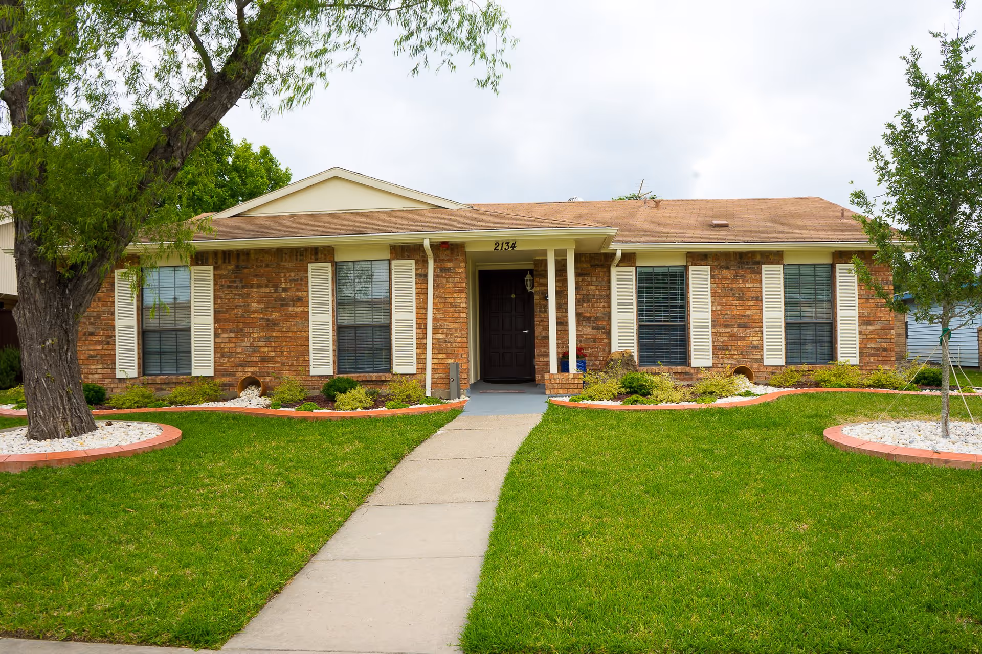 Front exterior view of a single-story brick house with a brown roof, white window shutters, and a central front door. The house is surrounded by a well-maintained green lawn, small landscaped garden beds with bushes, and two trees on either side of the walkway leading to the entrance.