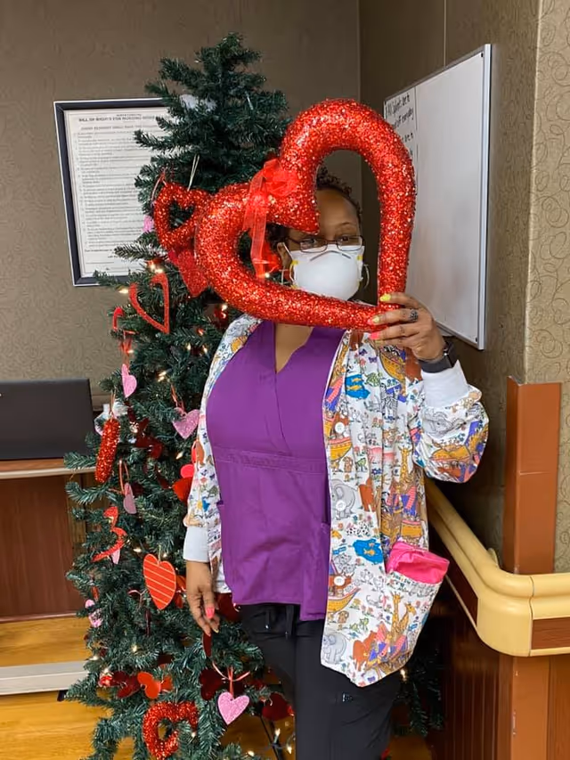 A person wearing a purple scrub top, a colorful patterned jacket, glasses, and a white face mask is standing indoors next to a decorated Christmas tree. The person is holding a large red glittery heart-shaped decoration in front of their face. The Christmas tree is adorned with red and pink heart ornaments and lights. There is a framed document on the wall and a whiteboard in the background.