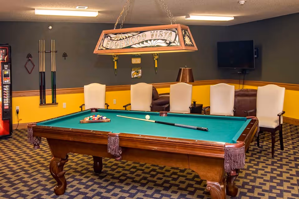 Interior game room with a green-felt pool table, cue sticks, chairs, a TV, vending machine, and a hanging 'Edgewood Vista' lamp.