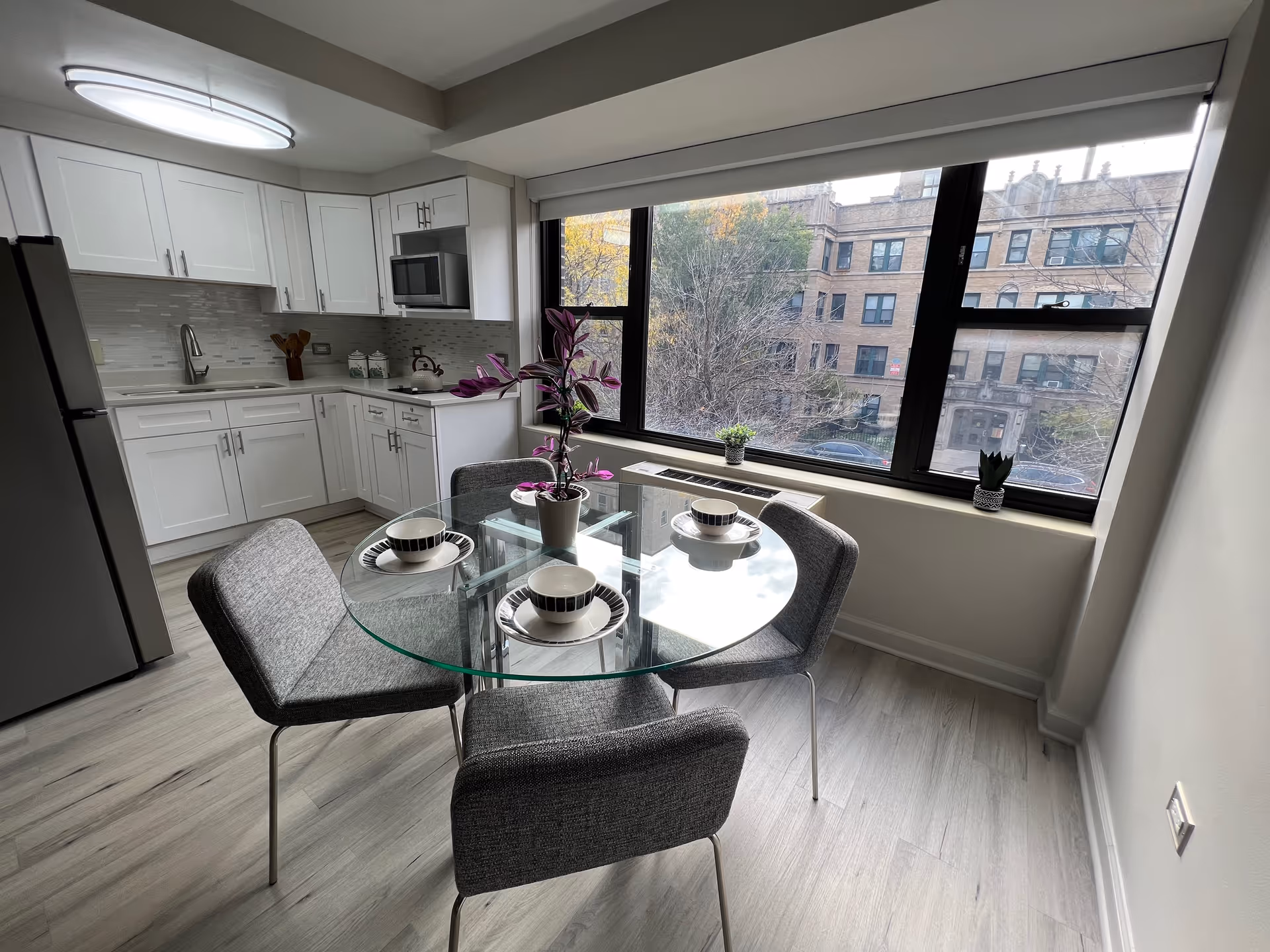 A modern kitchen and dining area with white cabinets, a stainless steel refrigerator, microwave, and a glass-top dining table set with four gray upholstered chairs. The table is set with four bowls and plates, and a potted plant is in the center. Large windows provide a view of an urban building and trees outside.